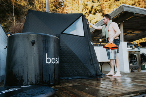 En kund vid sitt bastutält från bast med en bastuhink och handduk, med ett kallbadskar märkt bast i förgrunden