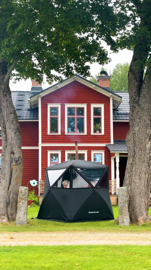 Svart bastutält från bast uppställt på gräsmattan framför ett rött hus, personer skymtar inuti, sommar