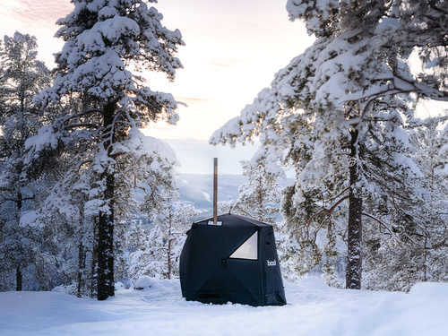Svart bastutält från bast med skorsten rest i djup snö bland snötyngda tallar, skogsklädd dal i bakgrunden en vinterkväll