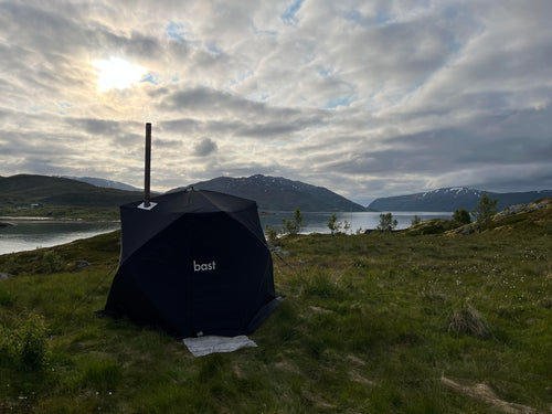 Svart bastutält från bast rest utomhus på fjällhed vid en fjord med snötäckta fjäll i bakgrunden, en fristående utomhusbastu
